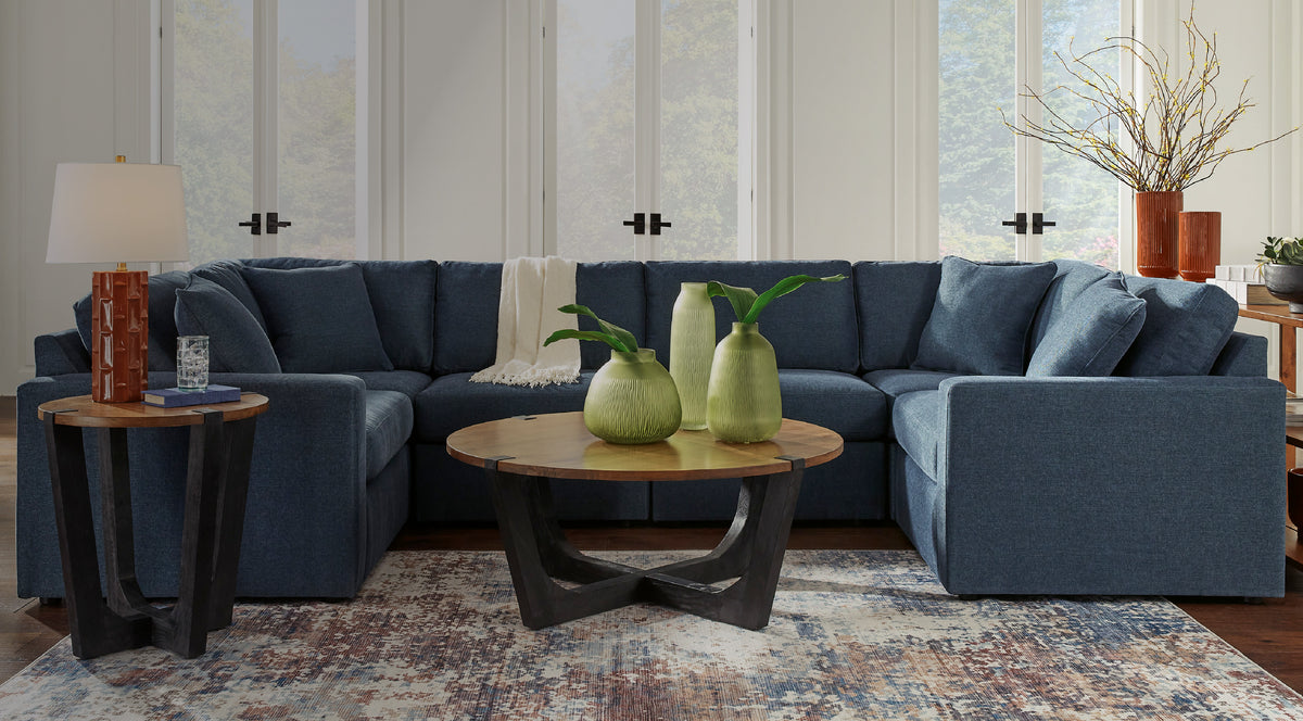 ModMax collection U-shaped modular sectional sofa in navy blue fabric with plush back cushions, paired with a round wood-top coffee table and matching end table on dark curved bases, styled on a multicolor distressed rug in a bright living room.